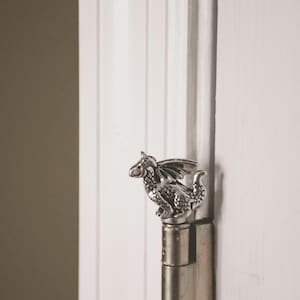 May include: A silver-colored dragon door hinge is mounted on a white door. The dragon has detailed wings and scales. The door is white with vertical paneling, and the background is a muted olive green.