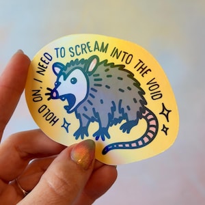 May include: A sticker featuring a cartoon opossum with its mouth open, surrounded by the text "I NEED TO SCREAM INTO THE VOID." The sticker has a yellow background with blue, gray, and pink accents.