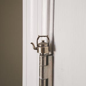 May include: A silver metal door hinge with a small, silver teapot attached to it.