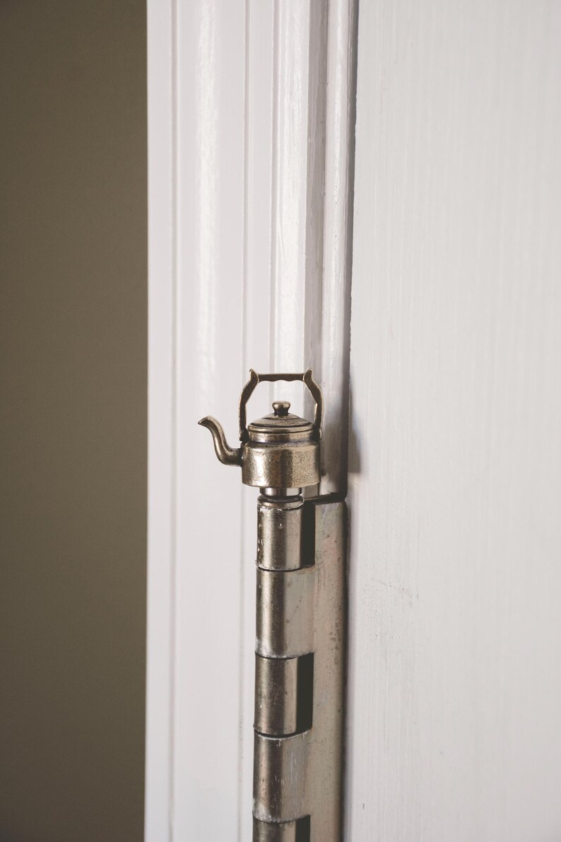 May include: A silver metal door hinge with a small, silver teapot attached to it.