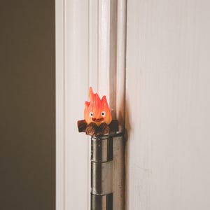 May include: A small orange and red figurine of a fire spirit sitting on a log, attached to a silver door hinge.