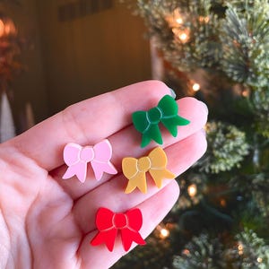 May include: A collection of colorful bow-shaped ornaments in pink, green, yellow, and red. The ornaments are small and appear to be made of a glossy material. The background includes a blurred Christmas tree.