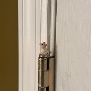 May include: A small, white ceramic mouse figurine with pink ears and nose accents, perched atop a metallic door hinge. The mouse is sculpted in a seated position, facing slightly to the left, and is attached to the top of the hinge pin.