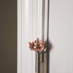 May include: A rose gold teapot-shaped door hinge. The hinge is attached to a white door.