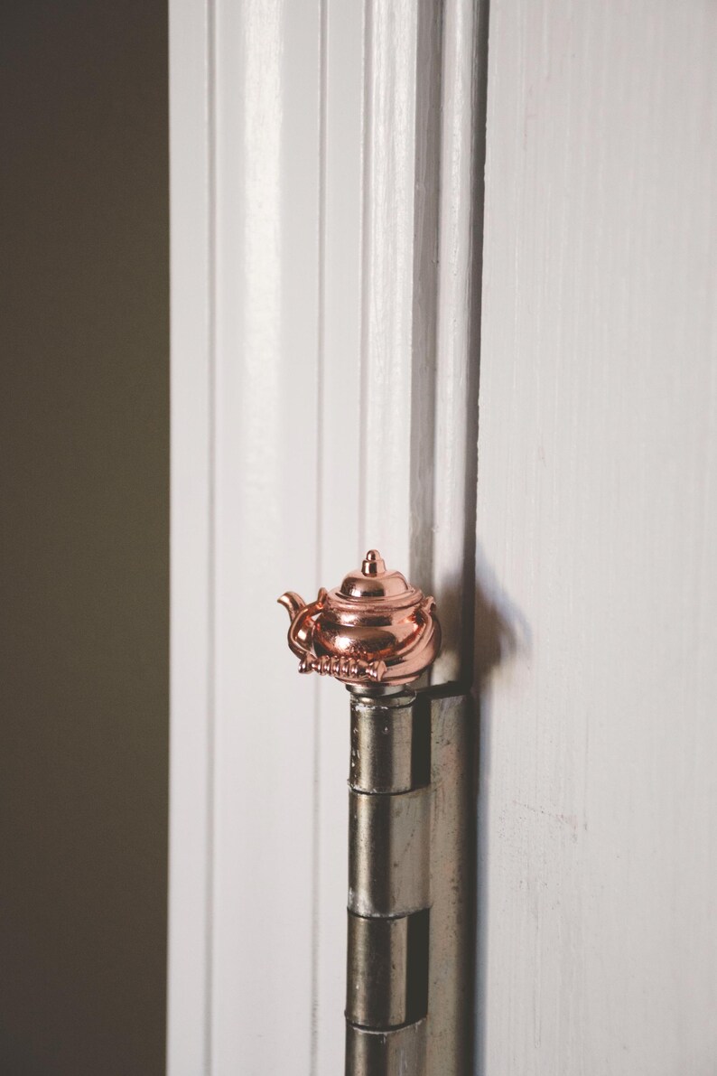 May include: A rose gold teapot-shaped door hinge. The hinge is attached to a white door.