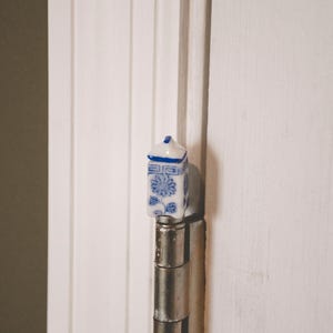 May include: A small, decorative porcelain cabinet knob with a blue and white floral design. The knob is rectangular with a small, pointed top and is mounted on a silver hinge. The background is a white door.
