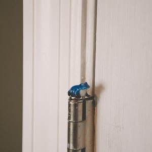 May include: A small, blue and white ceramic frog figurine perched on a metal door hinge. The hinge is attached to a white door frame. The frog has a glossy finish and is detailed with dark blue and white paint.