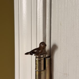 May include: A small, dark brown, polished stone seal figurine perched atop a metallic door hinge. The seal is sculpted with smooth, rounded features, including a defined head, flippers, and a tail, resting on a silver-toned metal cap.