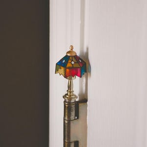 May include: A close-up of a door hinge with a decorative stained glass lamp finial. The lamp finial is made of glass and metal and features a colorful stained glass design. The hinge is made of metal and is attached to a white door.