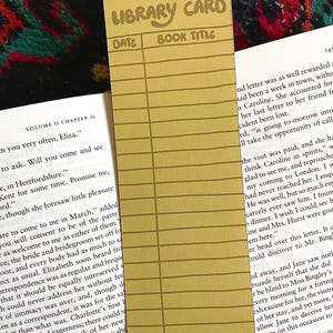 May include: A mustard-yellow library card bookmark with the words "LIBRARY CARD," "DATE," and "BOOK TITLE" printed on it. The bookmark has lines for entries and is placed on top of an open book.