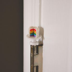 May include: A rainbow layered cake with white frosting on a small white cake stand. The cake is attached to a door hinge.