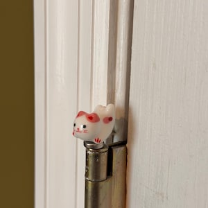 May include: A small, white ceramic cat figurine with pink ears and red facial details, including whiskers and a mouth, perched atop a metallic door hinge. The cat has a rounded body and is positioned horizontally on the top of the hinge pin.