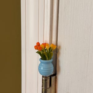 May include: A miniature light blue vase with horizontal ridges containing a small bouquet of orange and yellow tulip-like flowers. The vase is attached to the top of a metallic door hinge, positioned against a white vertical paneled door frame.