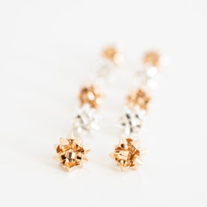 May include: A pair of gold and silver bow earrings. The earrings are made of multiple small bows that are linked together. The bows are in a staggered pattern, with the gold bows on one earring and the silver bows on the other.