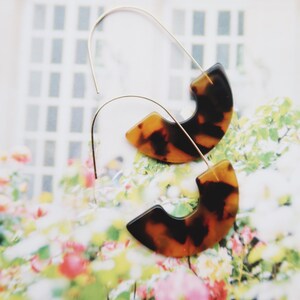 May include: A pair of earrings with a tortoiseshell pattern. The earrings have a gold-colored wire that curves into a semi-circle shape, with a tortoiseshell-patterned pendant. The background is a floral pattern.