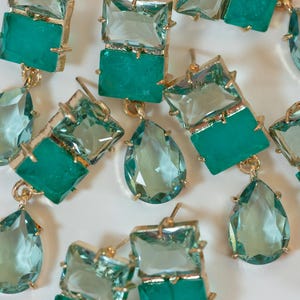 May include: Close-up of several pairs of earrings with gold-colored settings. Each earring features a combination of rectangular and teardrop-shaped, teal-colored gemstones. The earrings are arranged against a white background.