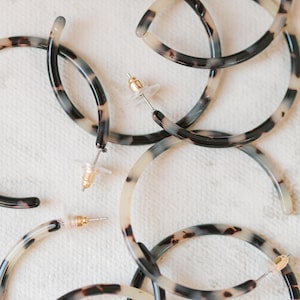 May include: A collection of tortoiseshell hoop earrings with gold-colored posts and clear backings. The earrings have a marbled pattern in shades of black, brown, and cream. The earrings are arranged on a white surface.