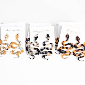 May include: Three pairs of snake-shaped earrings in different color patterns. The earrings are made of a tortoiseshell-like material. The earrings are displayed on white cards with the text "st. armonds Designs of Sarasota" printed on them.