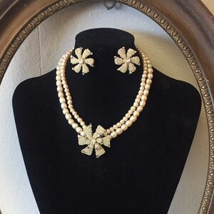 Può includere: Un set di gioielli in stile vintage con una collana di perle a doppia fila e orecchini abbinati. La collana e gli orecchini presentano un design floreale con dettagli scintillanti. Il set è esposto su un manichino nero all'interno di una cornice ovale dorata ornata.