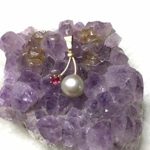Puede incluir: Un colgante de oro con una perla gris grande y redonda y una pequeña gema roja. El colgante se exhibe sobre un racimo de cristal de amatista púrpura, sobre un fondo blanco.