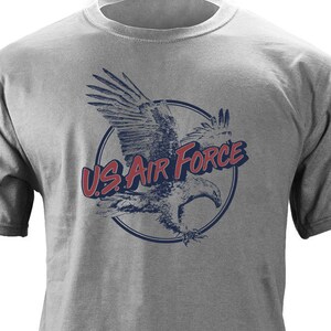May include: A gray t-shirt featuring a distressed graphic of an eagle inside a blue circle. The words "US AIR FORCE" are printed in red and blue, arching above the eagle. The shirt is made of a soft material.