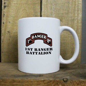 May include: White ceramic coffee mug with a black and orange graphic of the 1st Ranger Battalion insignia. The text "1st Ranger Bn" is printed in a black banner above the text "1st Ranger Battalion".