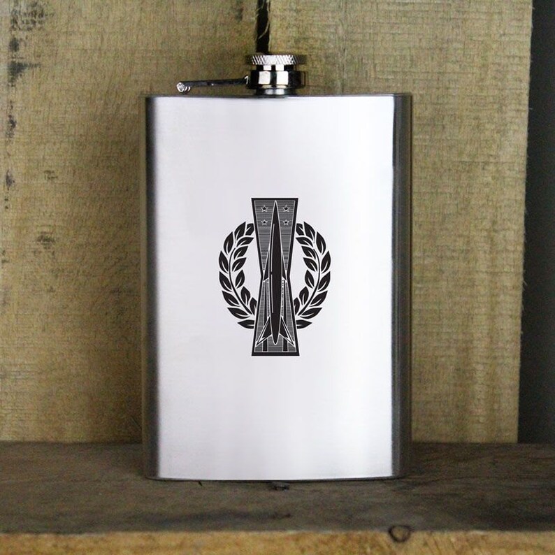 May include: A silver flask with a black and white graphic of a rocket ship inside a laurel wreath. The rocket ship has five stars above it.