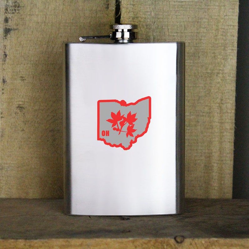 Puede incluir: Una petaca de plata con un contorno rojo del estado de Ohio y hojas de arce rojas dentro del contorno. La palabra "OH" est&aacute; en letras rojas debajo de las hojas.