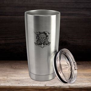 May include: A brushed stainless steel tumbler with a tapered cylindrical shape and a clear plastic lid. The front of the tumbler features a black engraved graphic of a sea creature with the text Honorable Shellback centered above it.