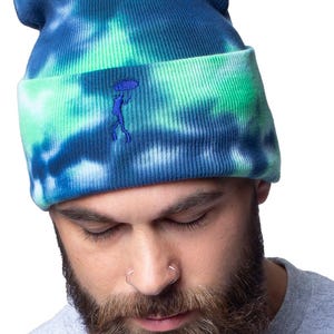 Puede incluir: Un gorro de punto azul y verde tie-dye con un pequeño diseño bordado en azul de una persona sosteniendo un paraguas.
