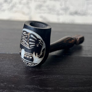 May include: A black wooden smoking pipe with a white elephant design carved into the bowl. The pipe is on a dark wooden surface.