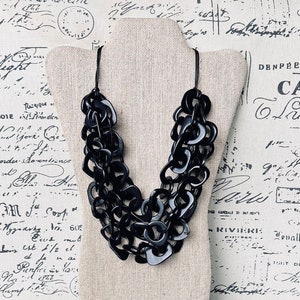 May include: A black chunky statement necklace made of multiple layers of large, rounded, interlocking links. The necklace is displayed on a fabric-covered mannequin bust.