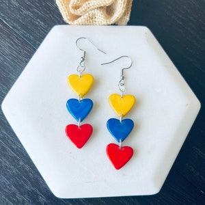 Puede incluir: Un par de pendientes colgantes con tres abalorios en forma de corazón. Los abalorios son amarillos, azules y rojos, y están dispuestos en una pila vertical. Los pendientes son plateados.