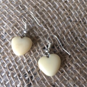 Puede incluir: Un par de pendientes en forma de corazón de color crema con ganchos plateados. Los pendientes se muestran sobre una superficie beige texturizada y tejida.