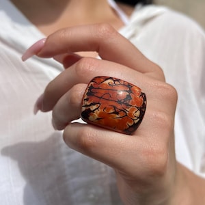 Peut inclure: Une bague carrée de grande taille avec un motif unique de tourbillons orange, marron et noirs. La bague est faite d'un matériau poli et son design est accrocheur. Cette bague est une pièce maîtresse, parfaite pour ajouter une touche artistique à n'importe quelle tenue.