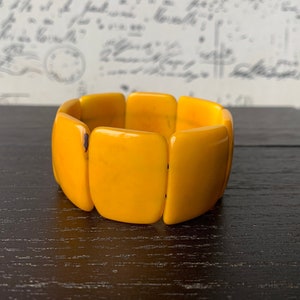 May include: A yellow plastic bracelet made of rectangular pieces. The bracelet is on a dark wood surface.