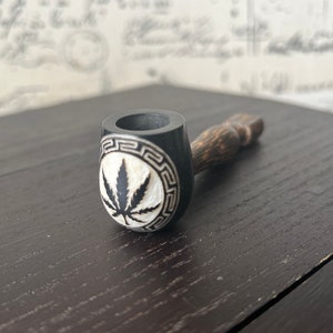 Op de afbeelding: Een zwarte en bruine houten pijp met een uitgesneden wit cannabisblad ontwerp. De pijp heeft een donkere afwerking met een decoratieve rand rond het blad. De steel is lichtbruin.