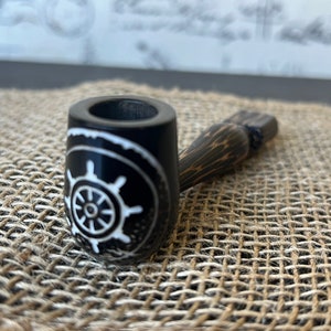 May include: A black wooden smoking pipe with a white ship's wheel design. The pipe is on a woven brown surface.