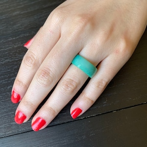 May include: A light teal-colored ring is worn on a hand with red nail polish. The ring is a solid band with a smooth, polished surface. The ring is a simple, elegant piece of jewelry.