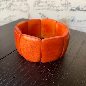 May include: Orange-colored, chunky bracelet with a marbled effect. The bracelet is made of several rectangular segments connected together. The jewelry is set against a dark surface, with a blurred background.