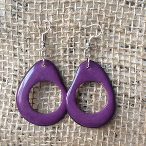 May include: A pair of purple teardrop-shaped earrings with a circular cutout. The earrings are suspended from silver-colored fish hook ear wires and a small chain. The earrings are displayed against a textured, neutral-colored background.