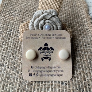 May include: A pair of cream-coloured tagua nut earrings on a brown card with the text "TAGUA STATEMENT JEWELRY Eco-friendly Fair trade Handmade Galapagos Tagua Galapagos Tagua.com E Galapagos Tagua.etsy.com fp @Galapagos Tagua"
