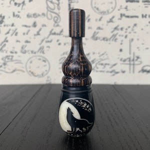 May include: A dark brown bottle stopper with a wolf howling at the moon design. The stopper is made of wood and has a decorative shape. The wolf image is in a white oval against a black background.