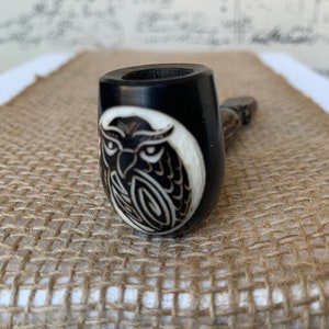 May include: A black wooden smoking pipe with an owl design carved into the bowl. The owl is white and black and has a detailed design.