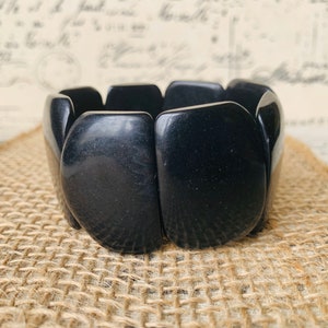 Pode incluir: Uma pulseira preta e grossa com um design elástico. A pulseira é feita de peças retangulares grandes e lisas. A joia é exibida em uma superfície texturizada bege.