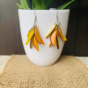 Puede incluir: Un par de pendientes colgantes con múltiples piezas de arcilla polimérica en forma de lágrima de color amarillo y naranja. Los pendientes cuelgan de ganchos plateados.