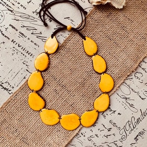 May include: A handmade necklace featuring large, teardrop-shaped yellow beads strung on a black cord. The necklace is displayed on a burlap-textured surface, with a small fabric flower in the background. The necklace is a statement piece.