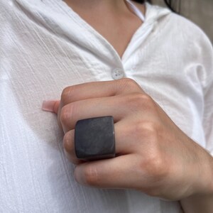 May include: A dark gray square ring. The ring is a solid, blocky shape, with a smooth, matte finish. The ring is worn on a finger, and the background is a white shirt.