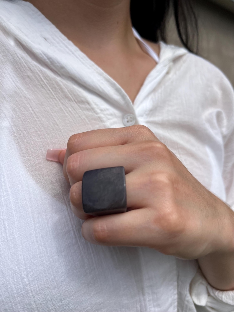 May include: A dark gray square ring. The ring is a solid, blocky shape, with a smooth, matte finish. The ring is worn on a finger, and the background is a white shirt.