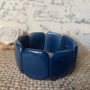Pode incluir: Um bracelete azul escuro feito de contas retangulares de cerâmica. O bracelete está sobre uma superfície de juta.